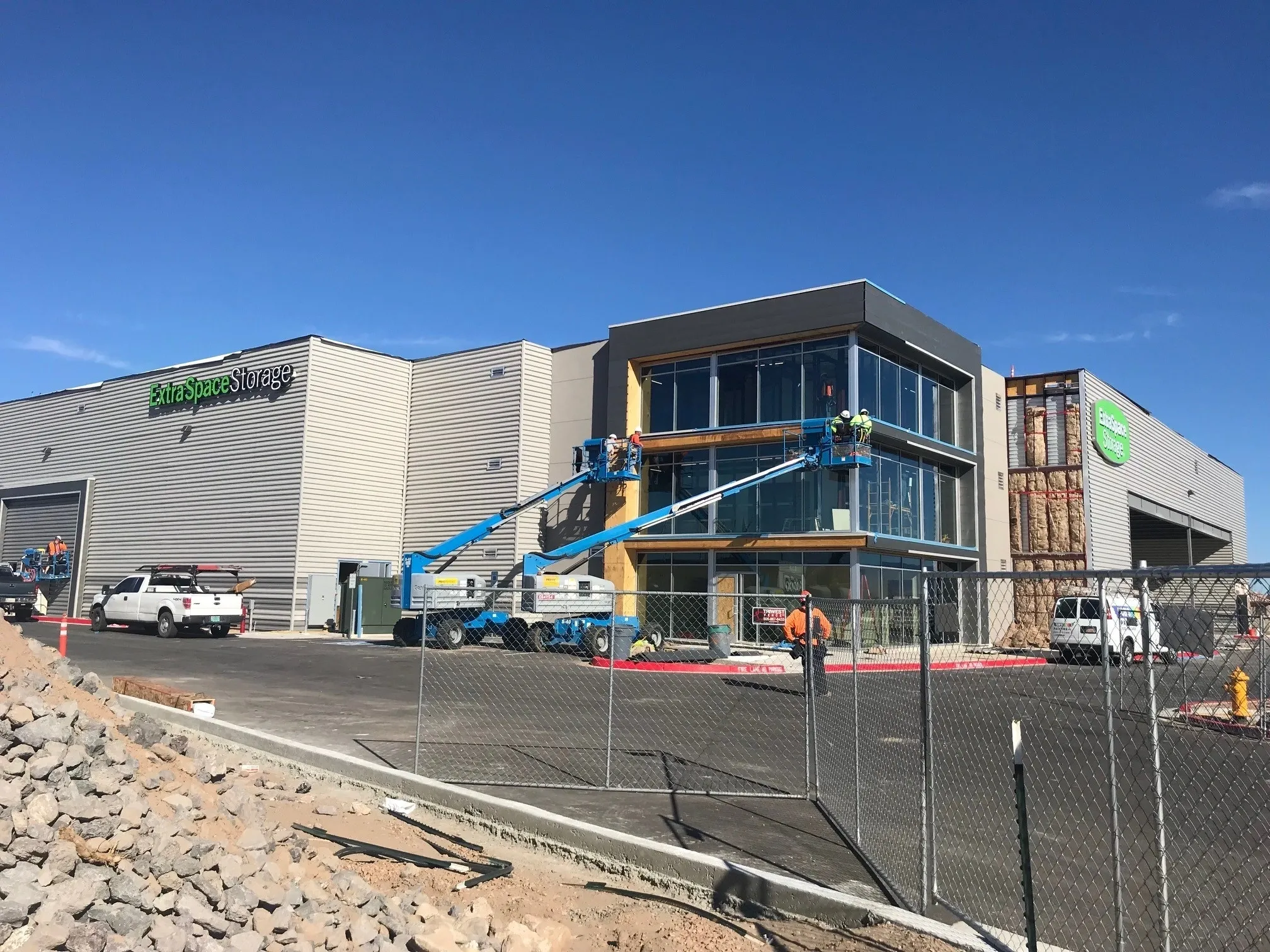 Construction workers on blue hydraulic lifts working on the exterior of a building, with two trucks and a fenced perimeter in the foreground under a clear blue sky.
